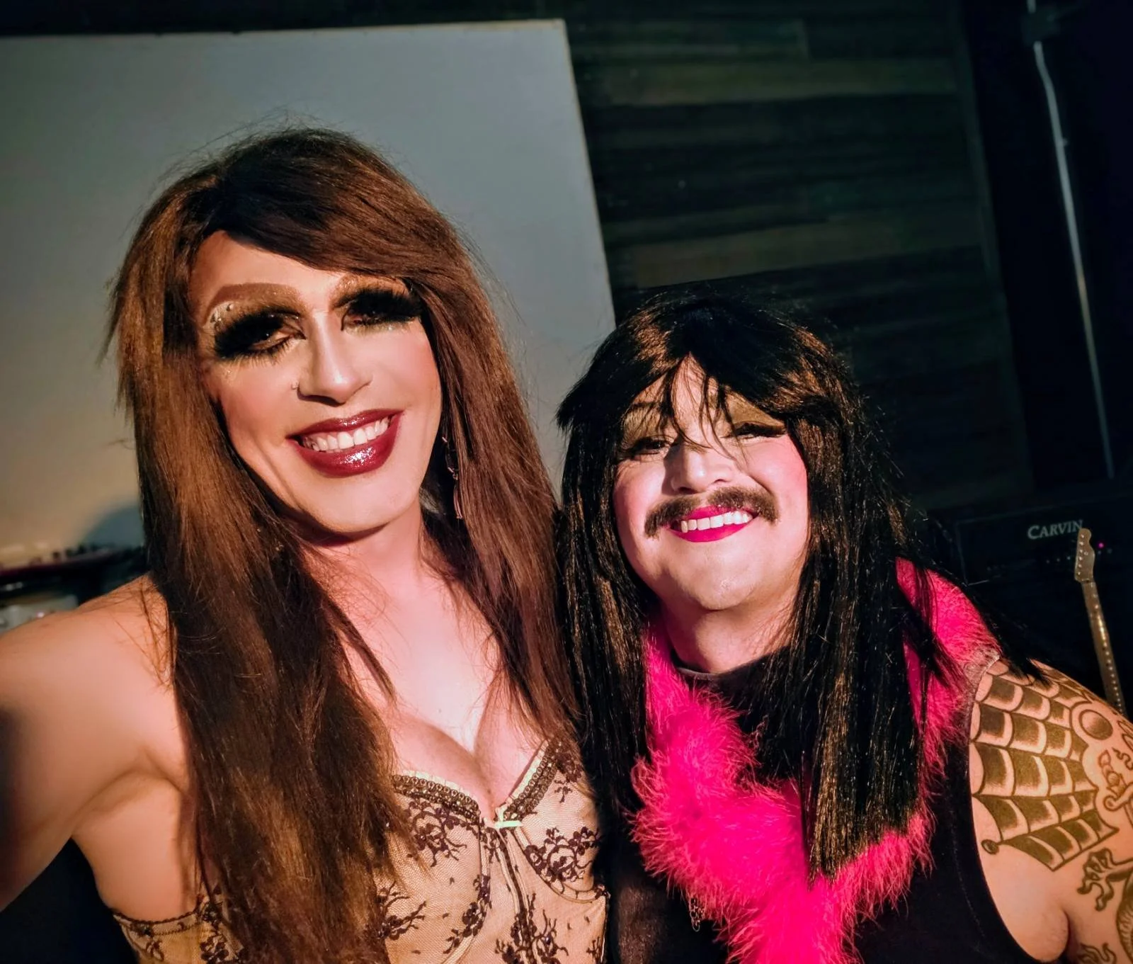 Two people dressed in drag, smiling at the camera. The person on the left has long brown hair, makeup, and a lace top, while the person on the right has black hair, makeup, a pink feathered boa, and tattoos on their arm.
