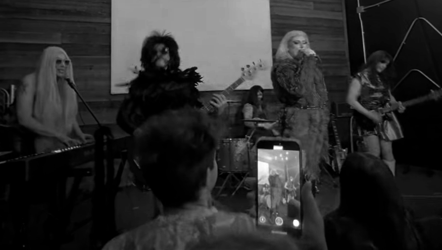 A band performing on stage with five members, including a keyboardist, guitarist, drummer, singer, and bassist, with an audience watching.