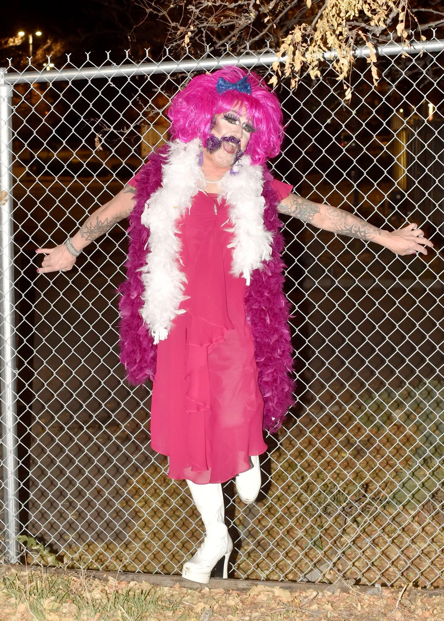 Person dressed in pink drag costume with bright pink wig and white boots, standing in front of a chain-link fence at night, arms outstretched.