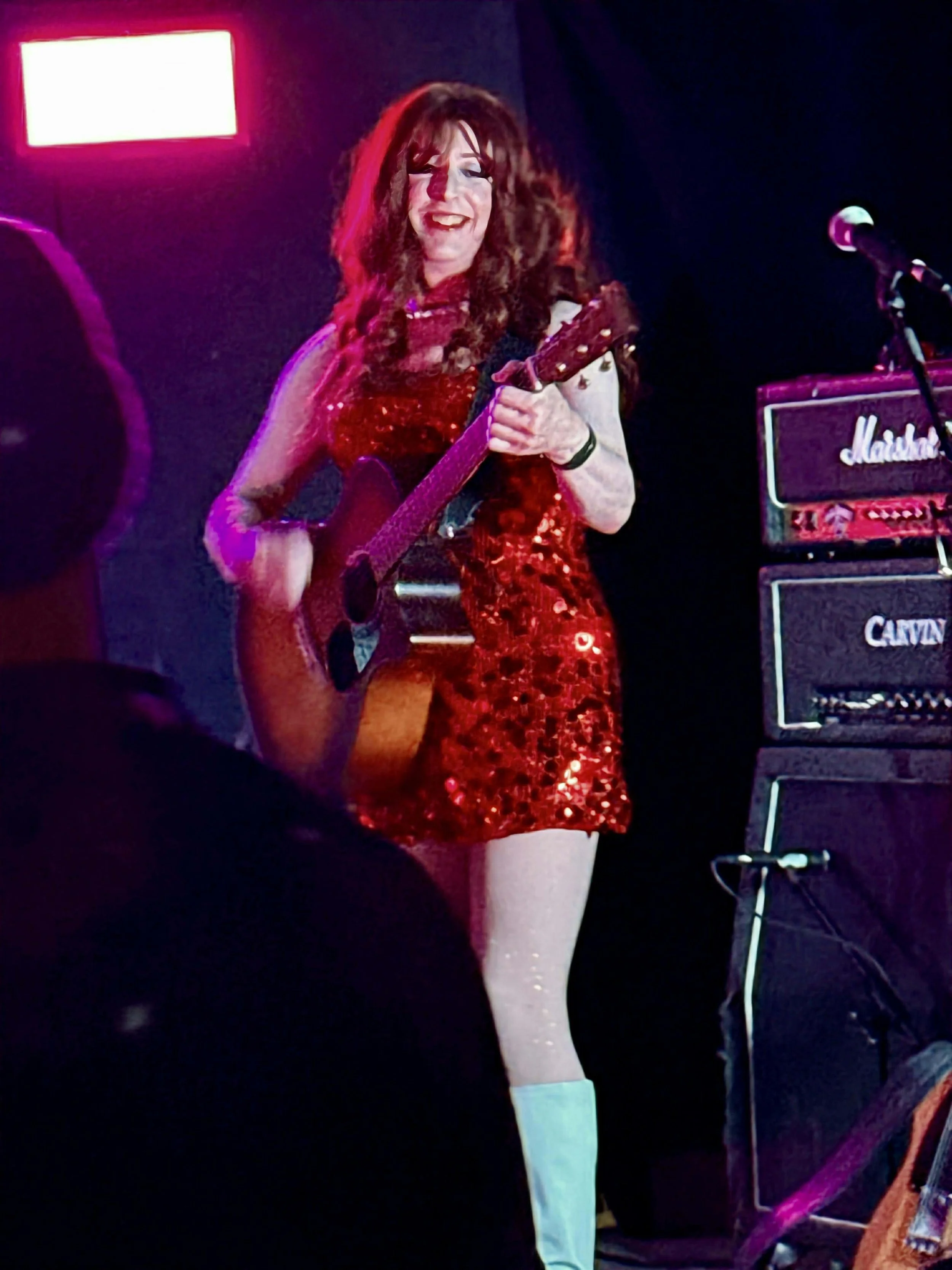 Woman with long red hair playing guitar on stage, wearing a red sequin dress and knee-high boots, with stage lights and amplifier in the background.