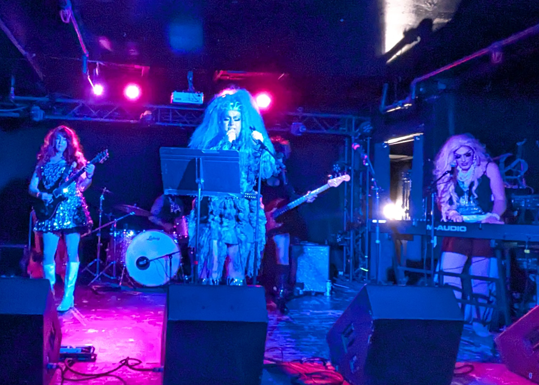 A drag band performing live on stage with four members playing various instruments under colorful stage lights.