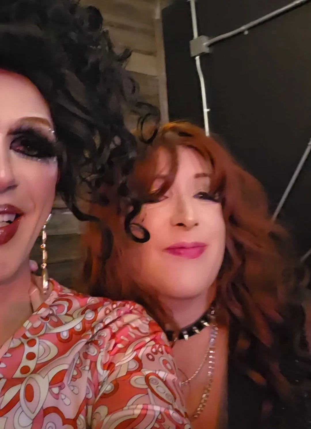 Two women with colorful hair and makeup taking a selfie at a social event, one with black curly hair and bold makeup, the other with red hair and soft makeup, wearing colorful patterned clothing and jewelry.