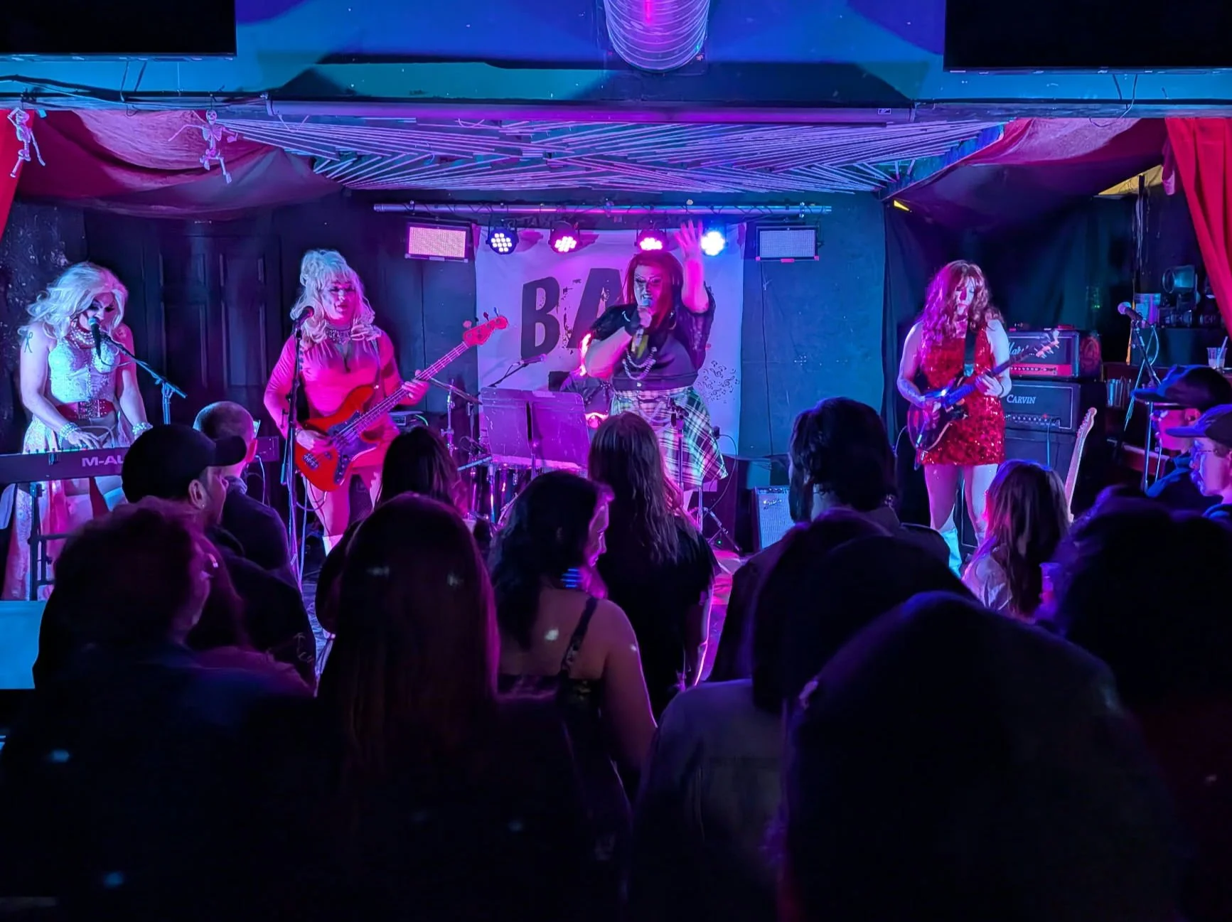 A live band performance by CoverGurls on stage at a venue with colorful lights, five drag queen singers and musicians, and an audience watching.