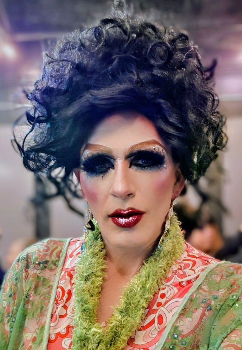 Close-up of a person with elaborate makeup, curly black hair styled in an updo, wearing a colorful patterned outfit with a green fuzzy scarf, and earrings.