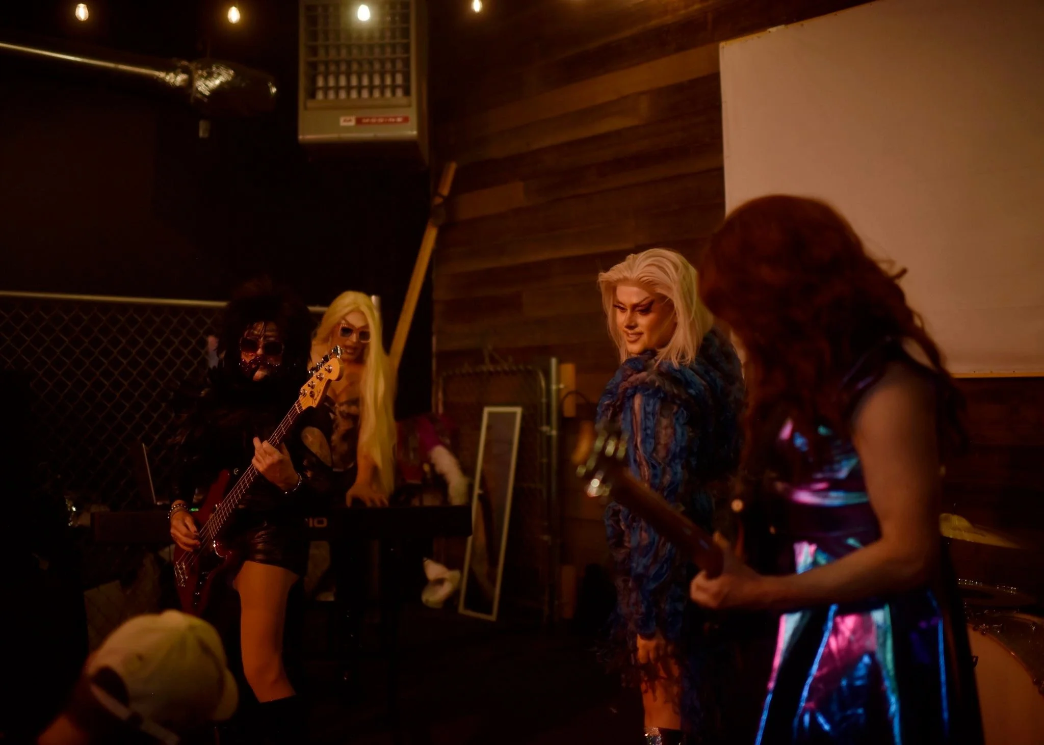 Four drag performers in a dimly lit room, one playing an electric guitar and another holding a bass guitar, with a keyboard and a mirror in the background, all dressed in edgy, colorful outfits.