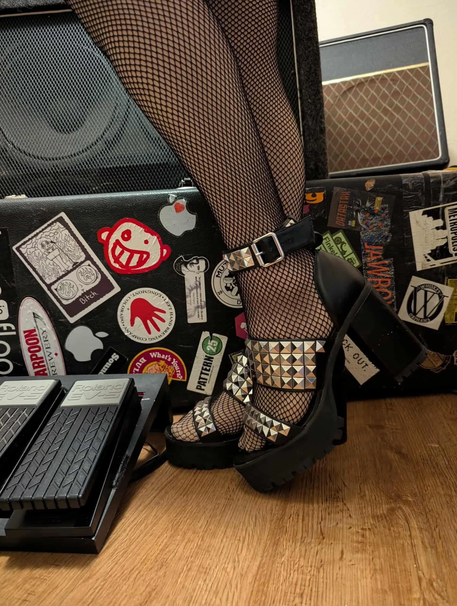 Close-up of a person's feet wearing black fishnet stockings and black platform sandals with pyramid-shaped studs, standing on a wooden floor near a black equipment case and a guitar amplifier.