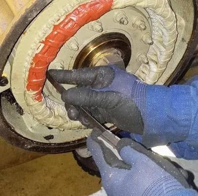 Pessoa realizando manutenção ou inspeção em uma peça mecânica com ferramentas, usando luvas de proteção.