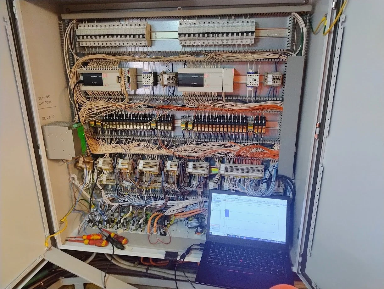 Quadro de painel elétrico com componentes e cabos, com um laptop na parte inferior exibindo um gráfico.