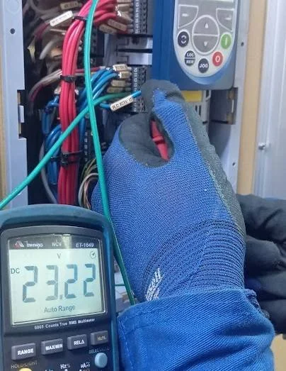 Pessoa usando uma luva está medindo a tensão de um equipamento elétrico com um multímetro, que mostra 23.22 volts.