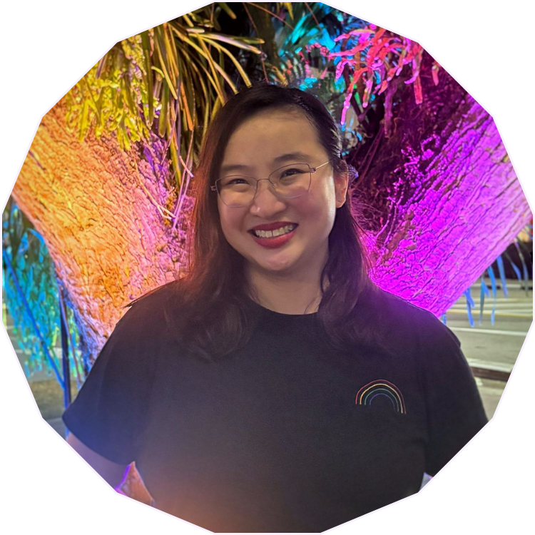 A smiling woman with glasses and dark hair standing in front of a tree illuminated with colorful rainbow lights.