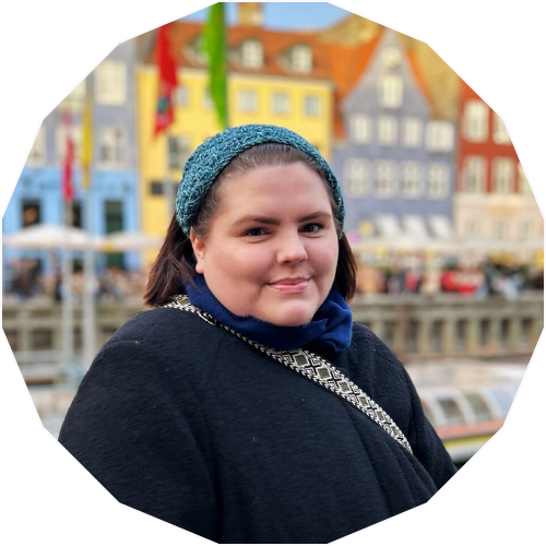A young woman wearing a blue knit hat and a black coat, standing outdoors with colorful buildings and a canal behind her.