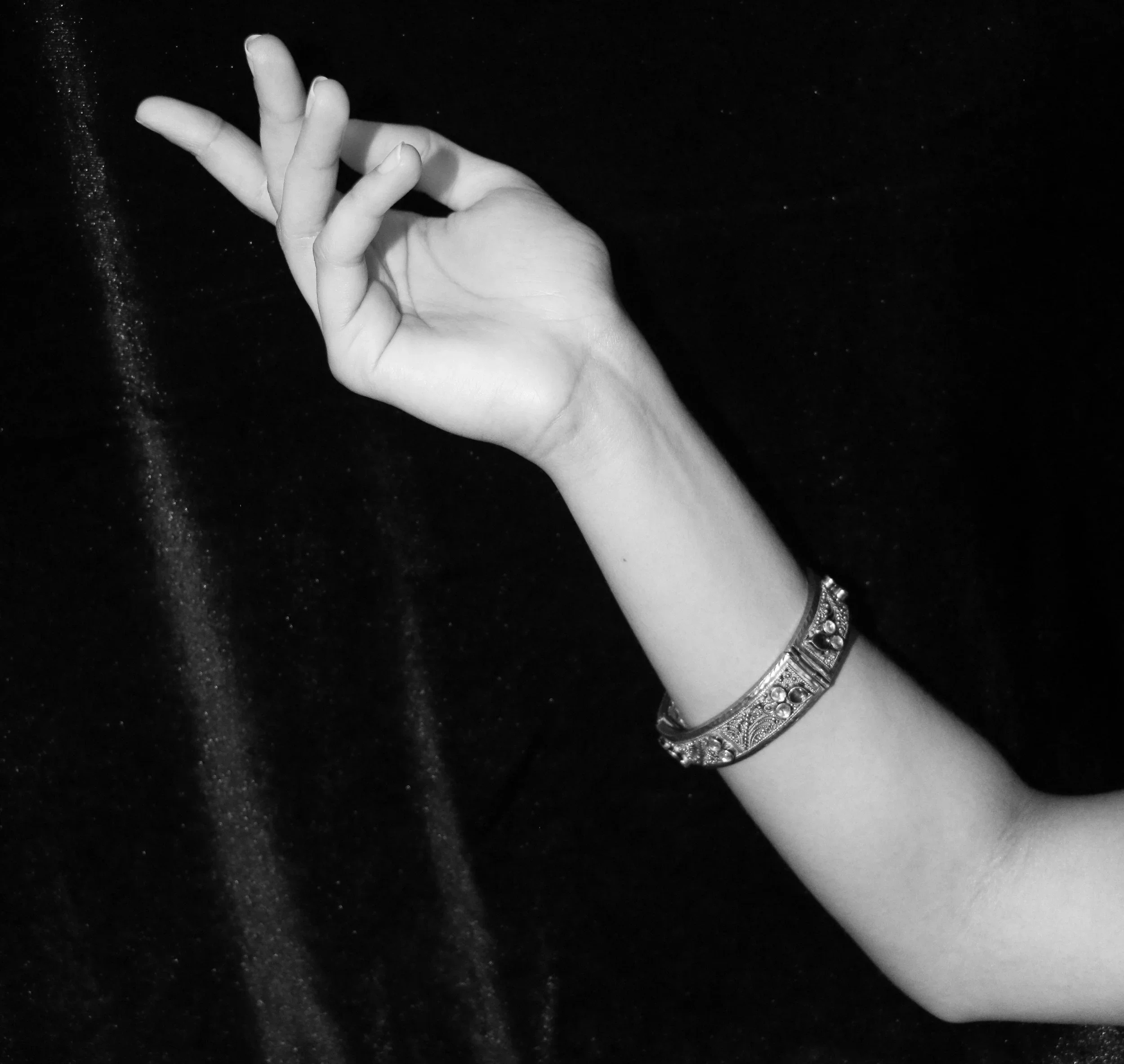 Black and white photo of a left hand with fingers forming an okay gesture, wearing a decorative bracelet on the wrist, against a dark, textured background.