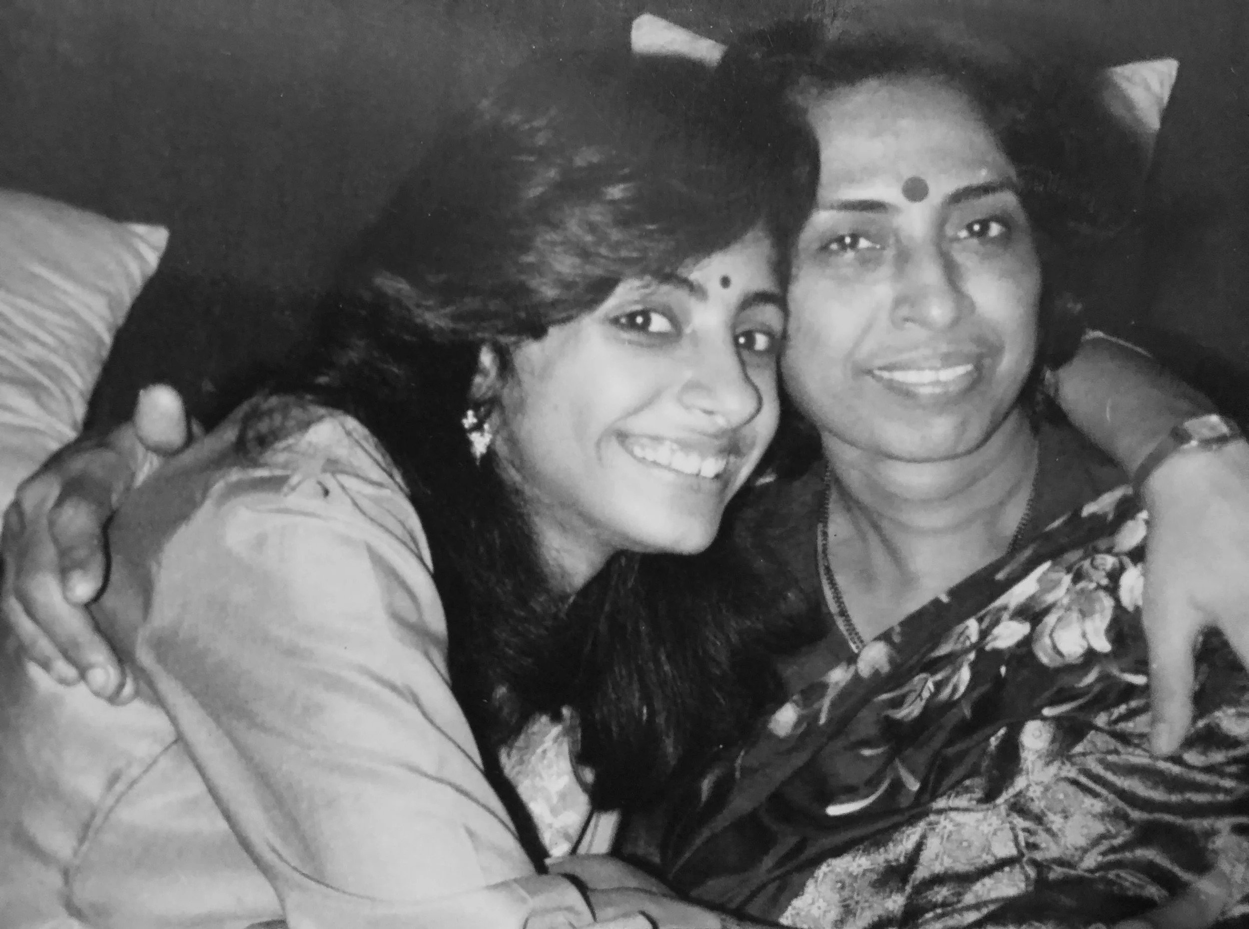 Two women sitting close together, smiling, with their arms around each other, in a black-and-white photo.