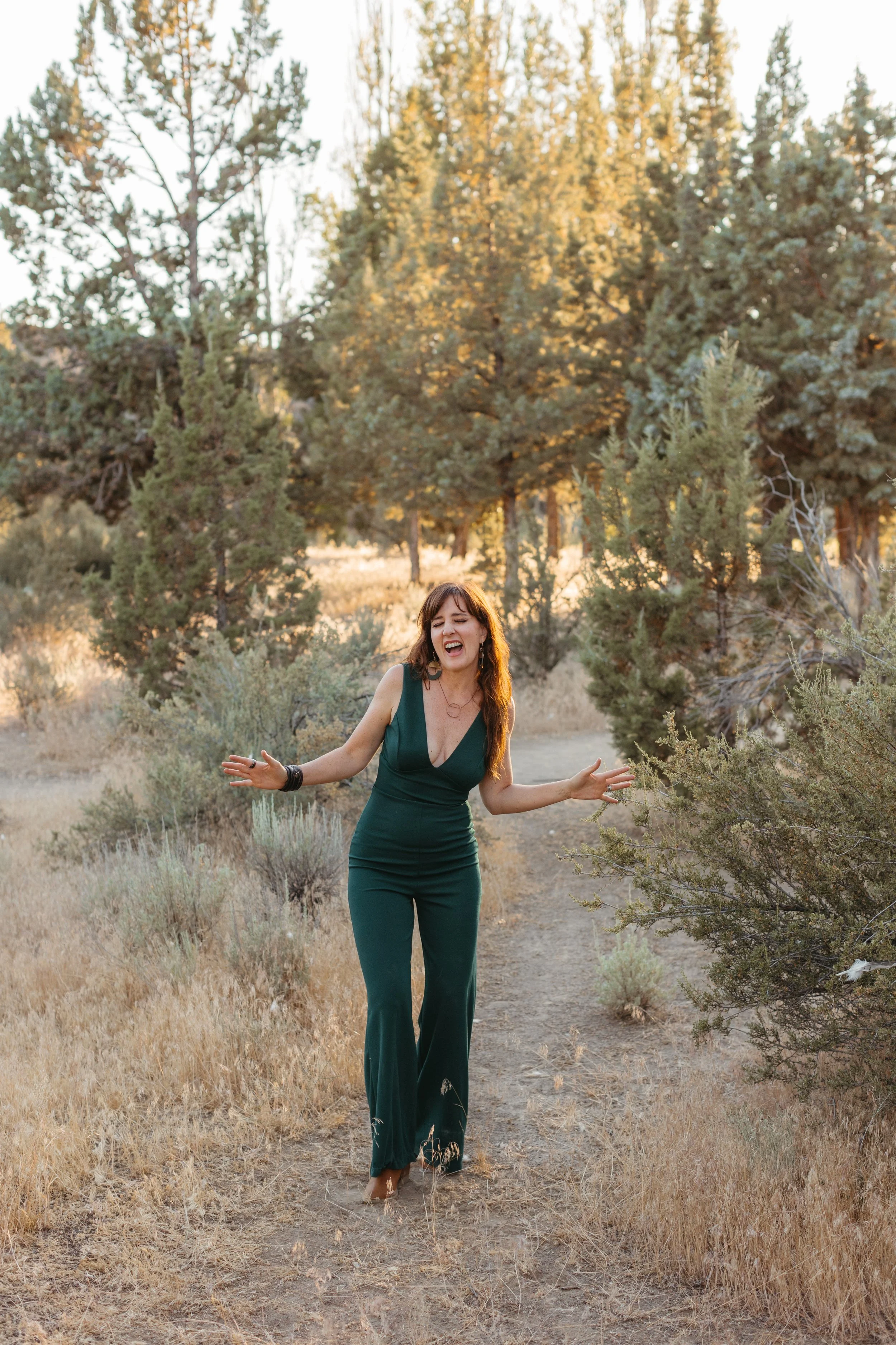 A woman in a green jumpsuit walking through a dry, wooded area during sunset, smiling and gesturing with her arms.