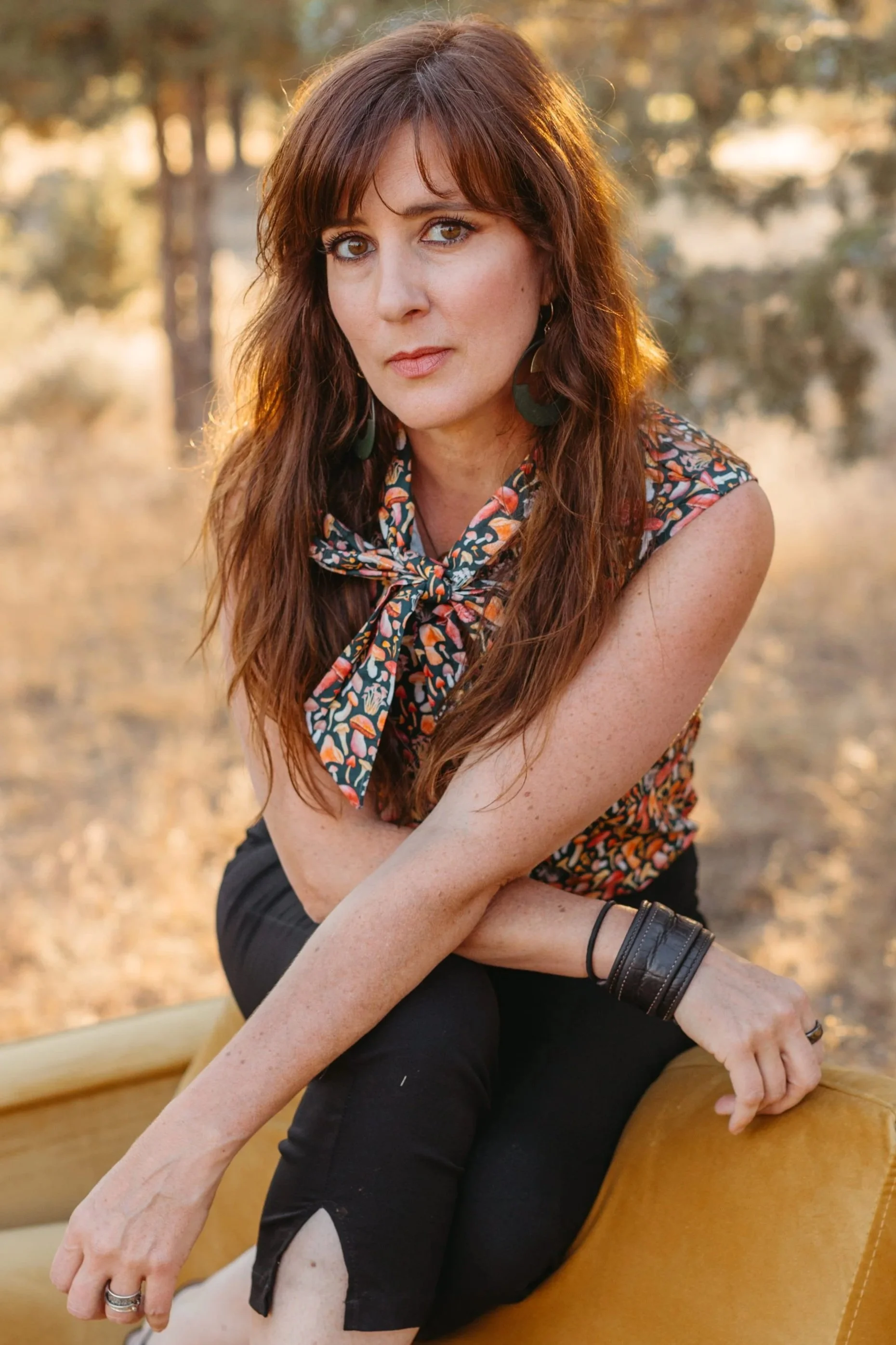 A woman with long brown hair and brown eyes sitting outdoors on a yellow couch, wearing a sleeveless floral top with a bow tie and black pants, during golden hour with trees and sunlight in the background.