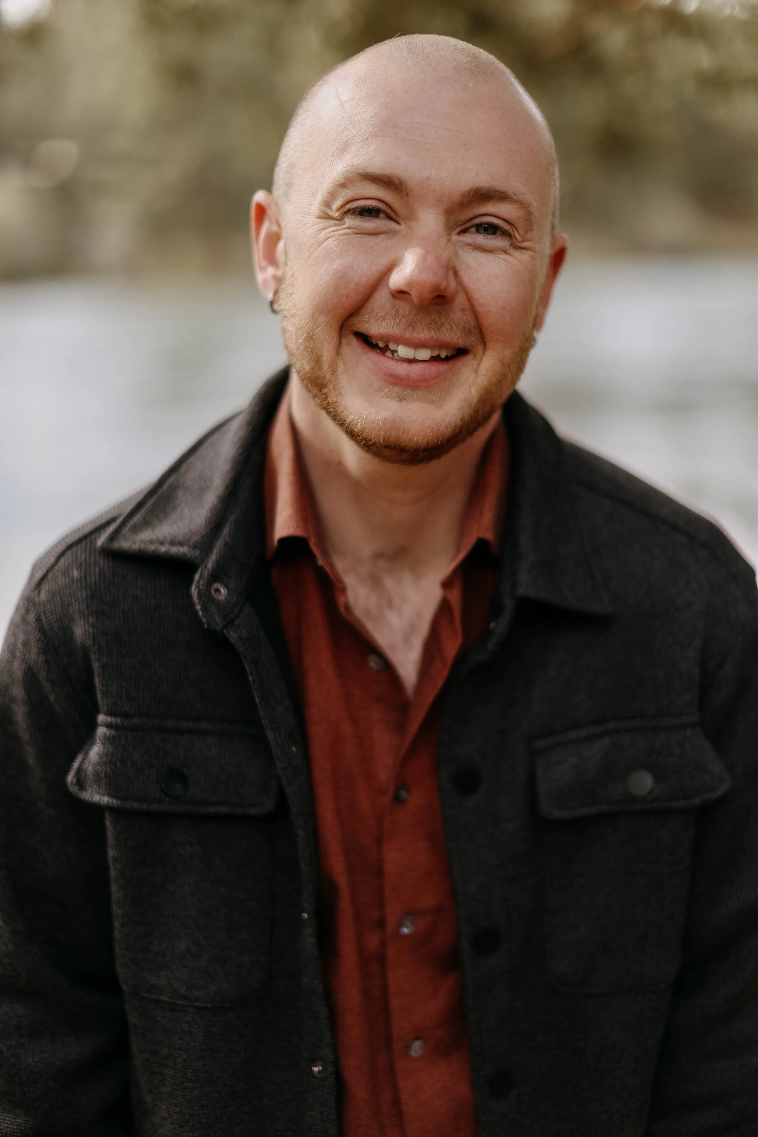 A smiling man with a shaved head, beard, and earrings, wearing a dark jacket over a rust-colored shirt, outdoors near water with blurred trees in the background.