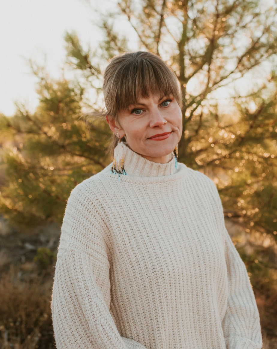 A woman with short brown hair wearing a white knitted sweater and colorful earrings outdoors during sunset, with trees and a softly illuminated sky in the background.