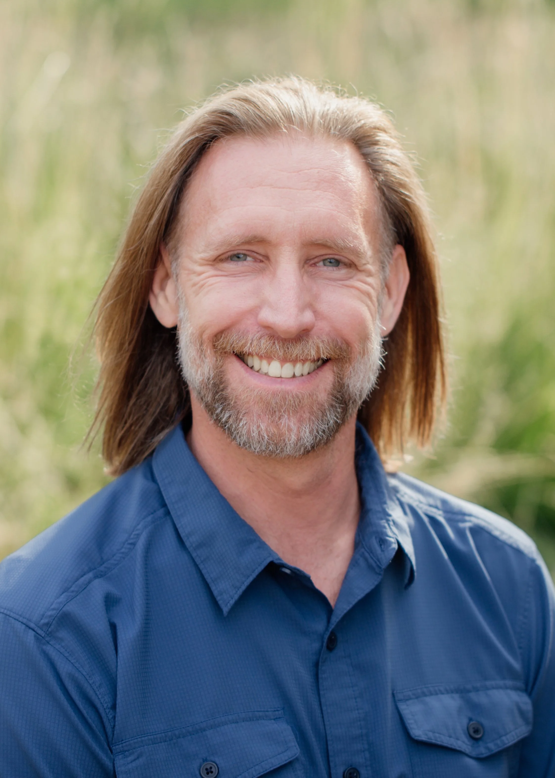 A smiling man with long brown hair, a beard, and blue eyes, wearing a blue button-up shirt, outdoors with greenery in the background.