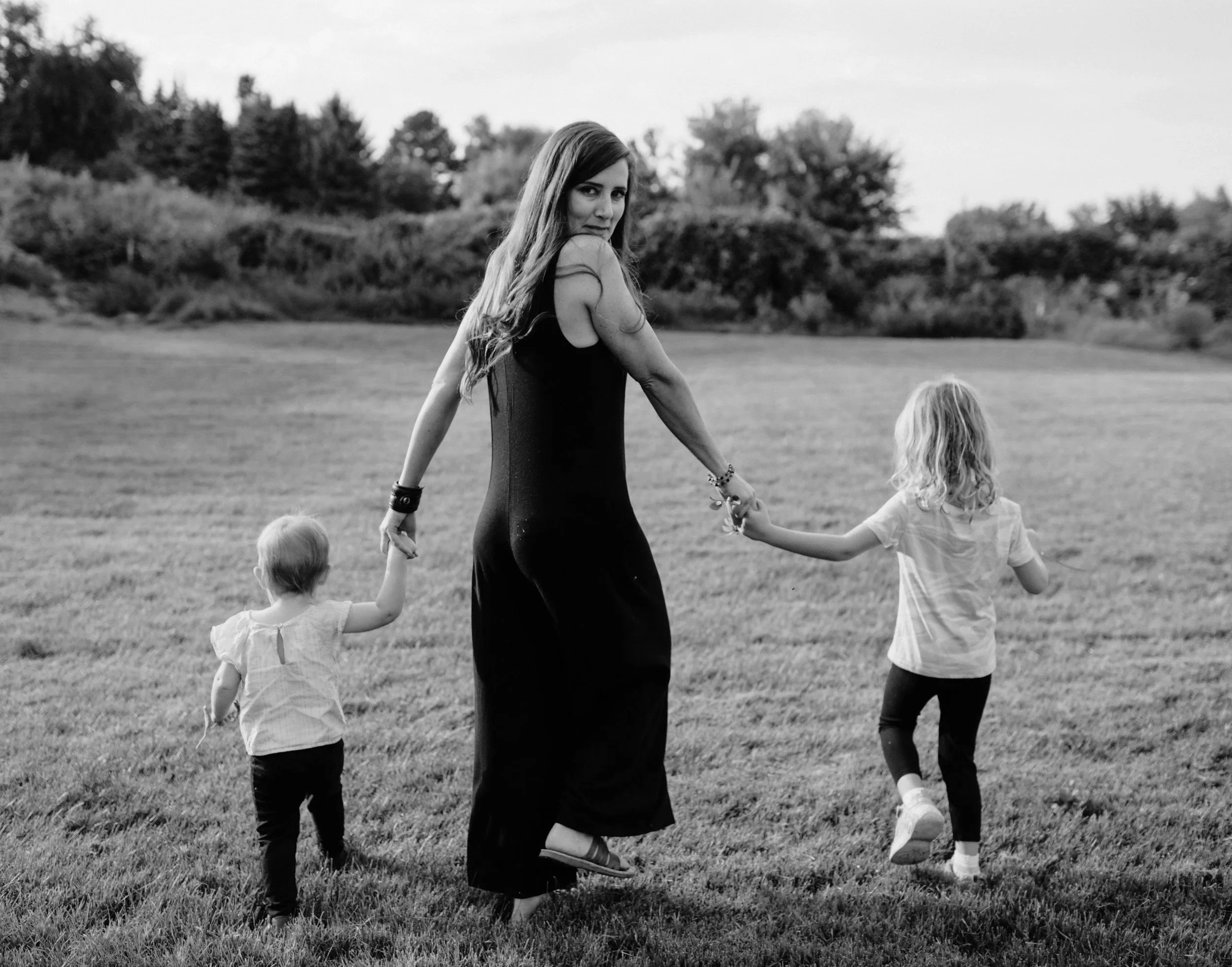 A woman walking on a grassy field holding hands with two young children, one on each side, with a background of trees.