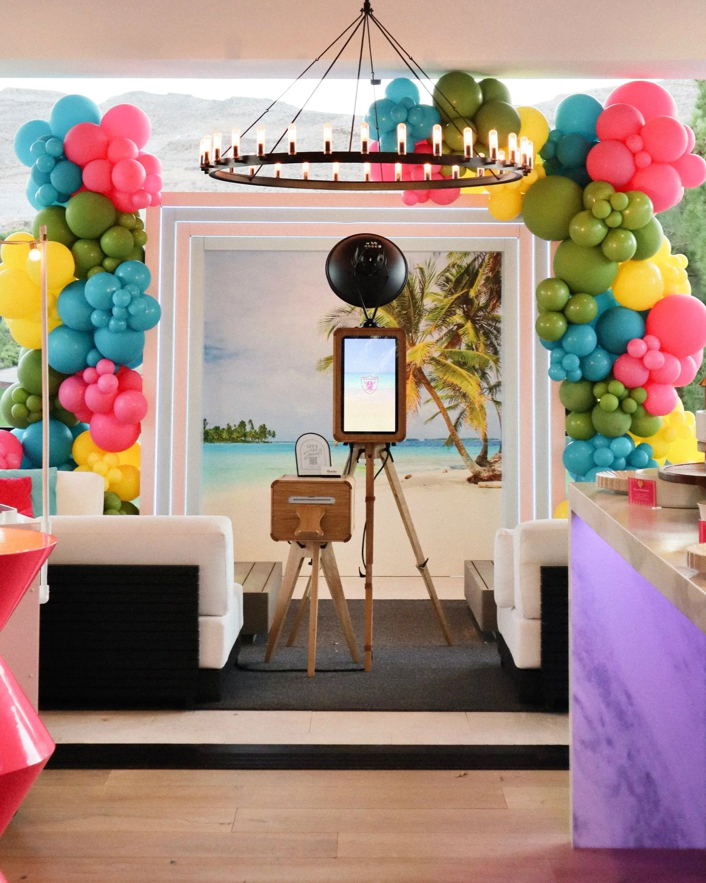 Decorative photo booth in Las Vegas with tropical beach background, balloon arch with pink, blue, green, and yellow balloons, tripod with photo booth equipment, and chandelier with small lights.