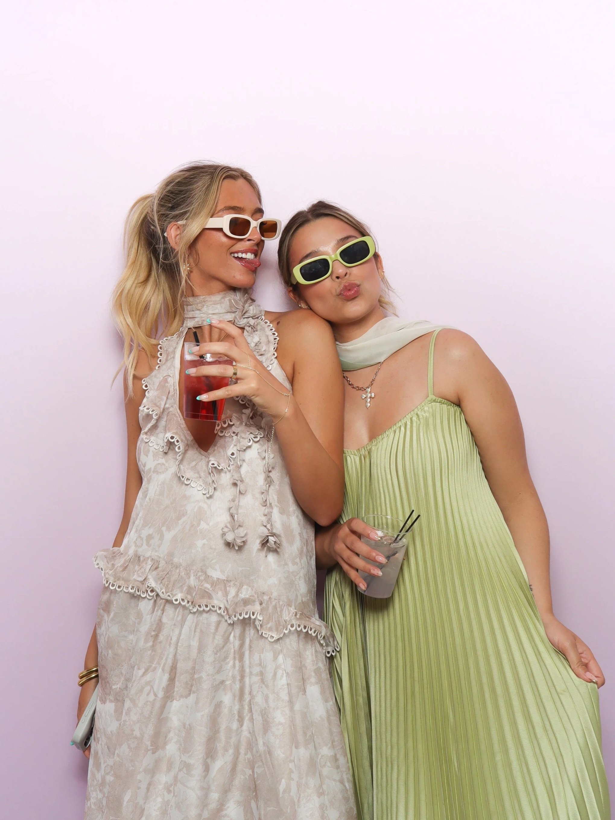 Two women standing close together, wearing stylish sunglasses and light-colored summer dresses, smiling and enjoying photo booth at a wedding in Las Vegas
