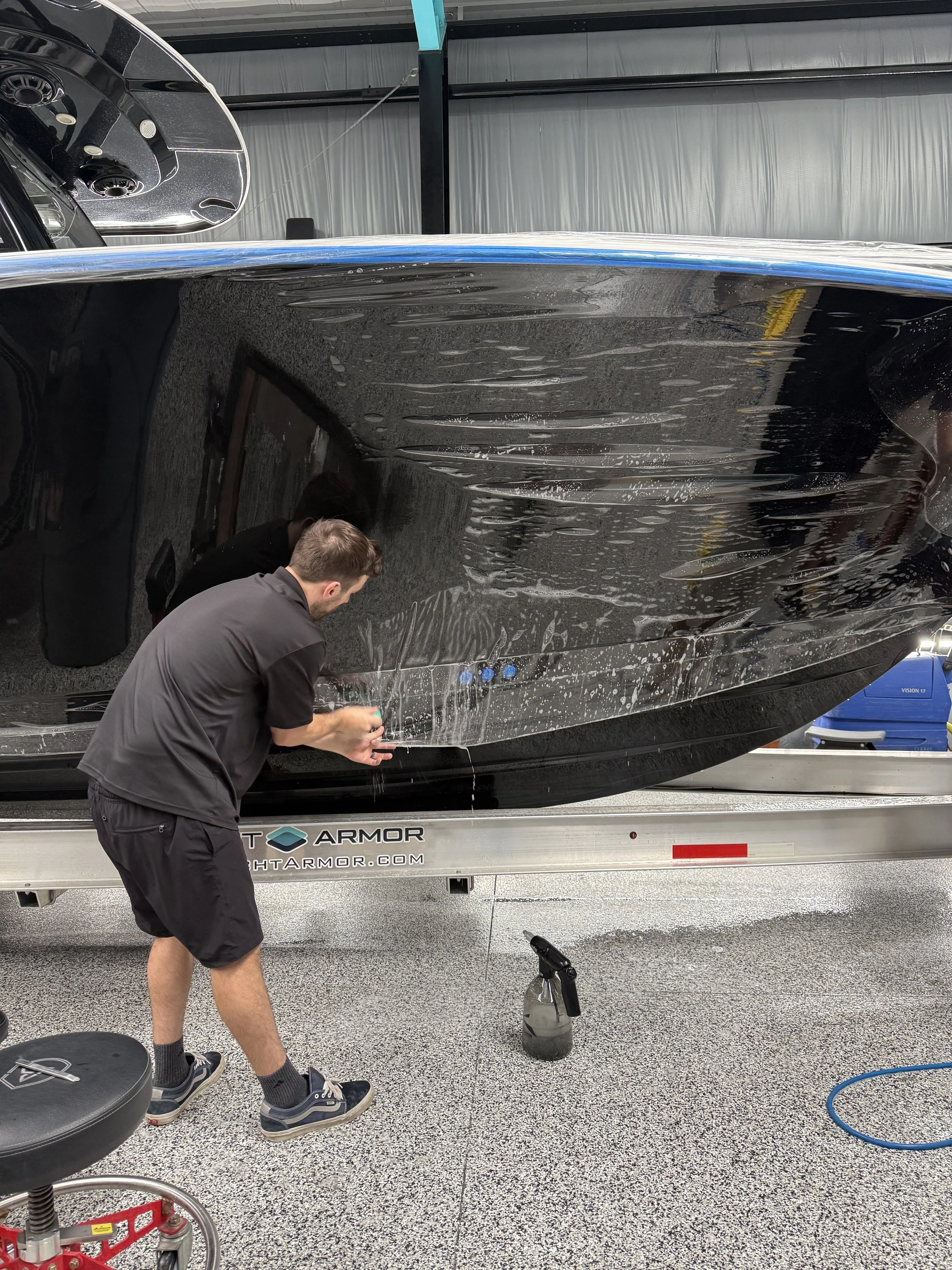 A man cleaning the hull of a large black boat inside a garage or workshop.