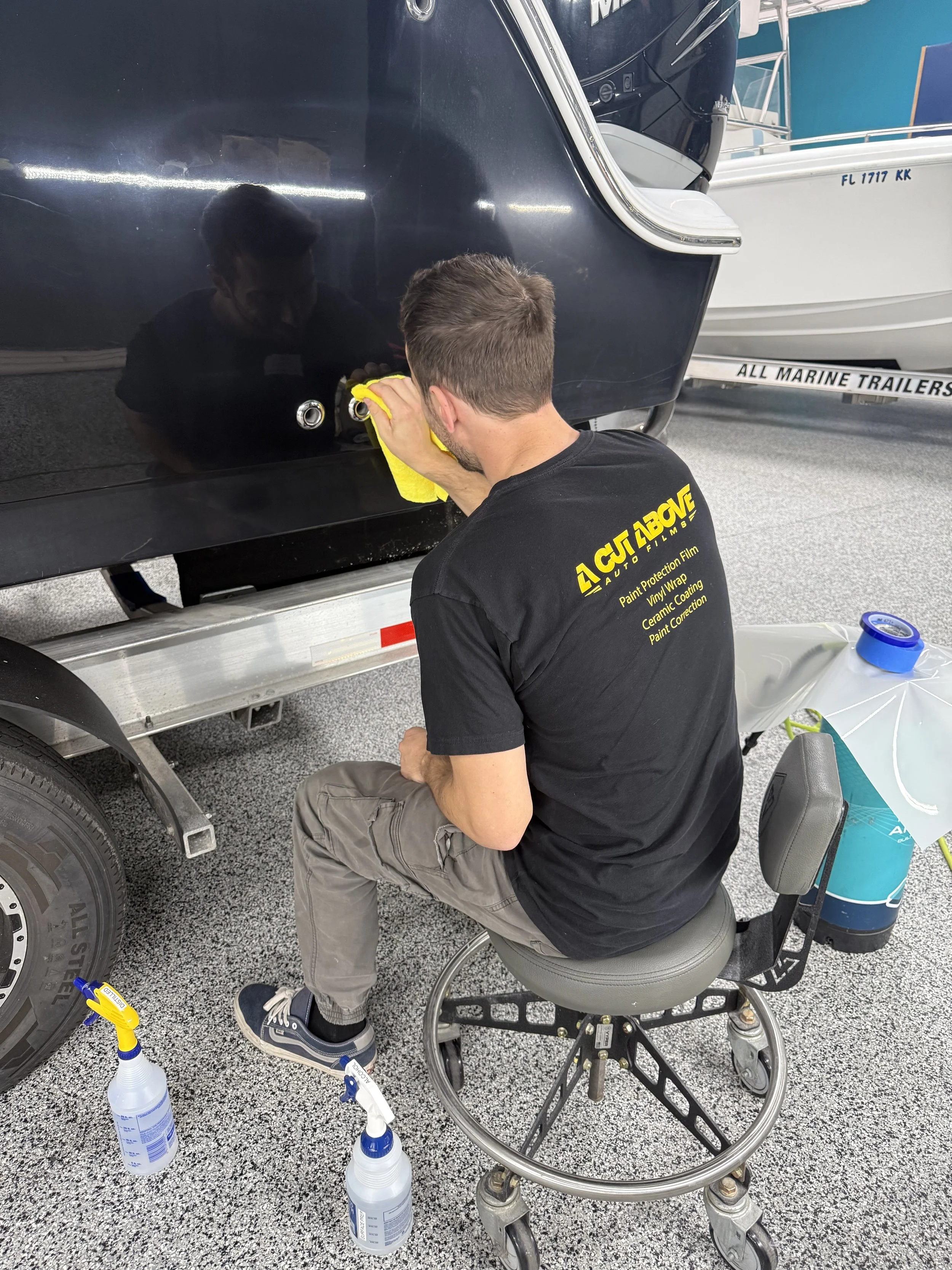 A man cleaning or polishing a black vehicle with a yellow cloth in a garage, with boats in the background and bottles of cleaning supplies on the floor.