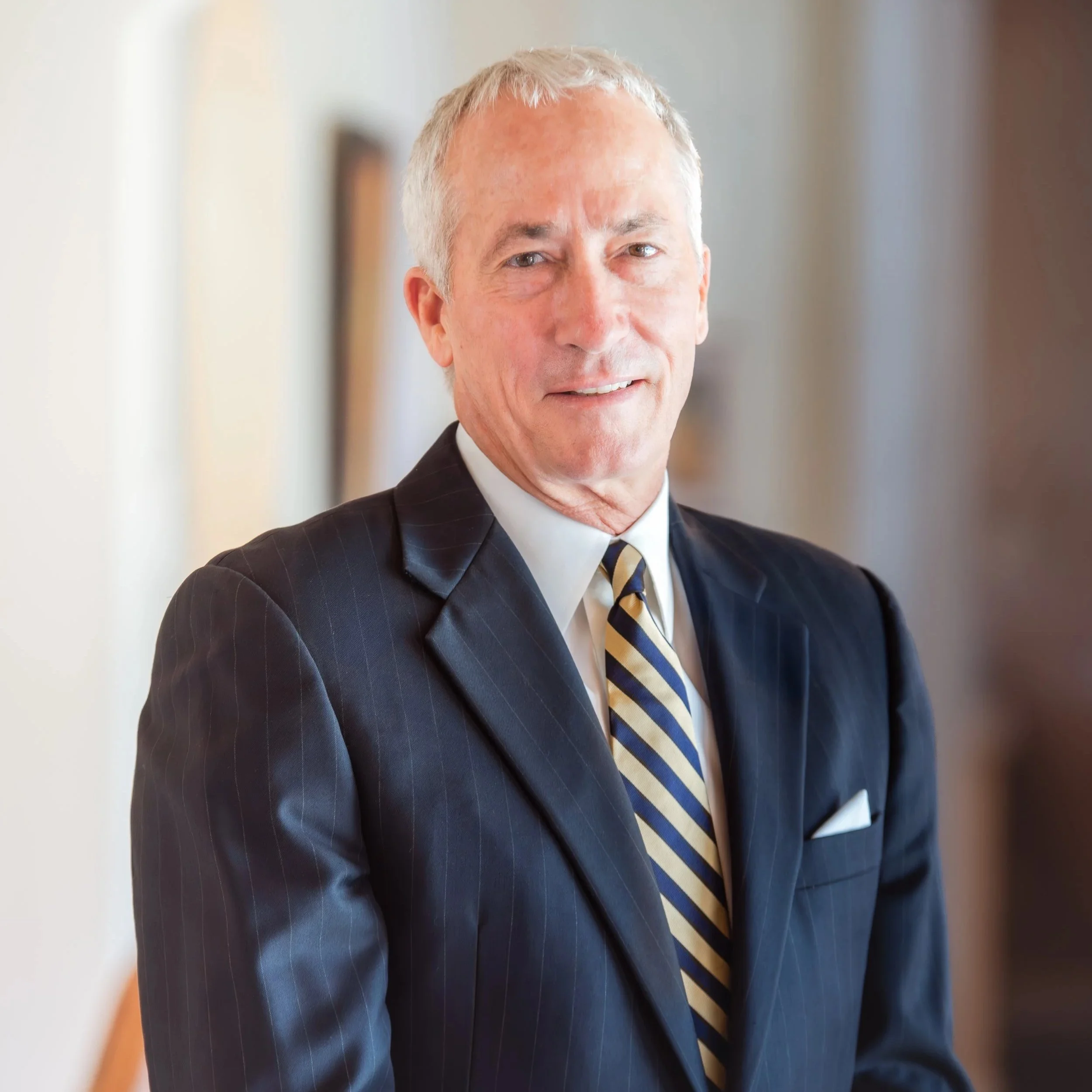 A mature man in a dark pinstripe suit with a white shirt and a striped tie, standing indoors with a blurred background, smiling slightly, facing the camera.