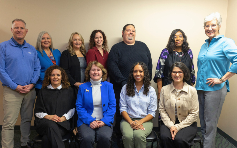 A group of ten adults, five women and five men, posing together in an indoor setting. They are dressed in casual and business casual attire, standing and sitting in two rows, smiling at the camera.