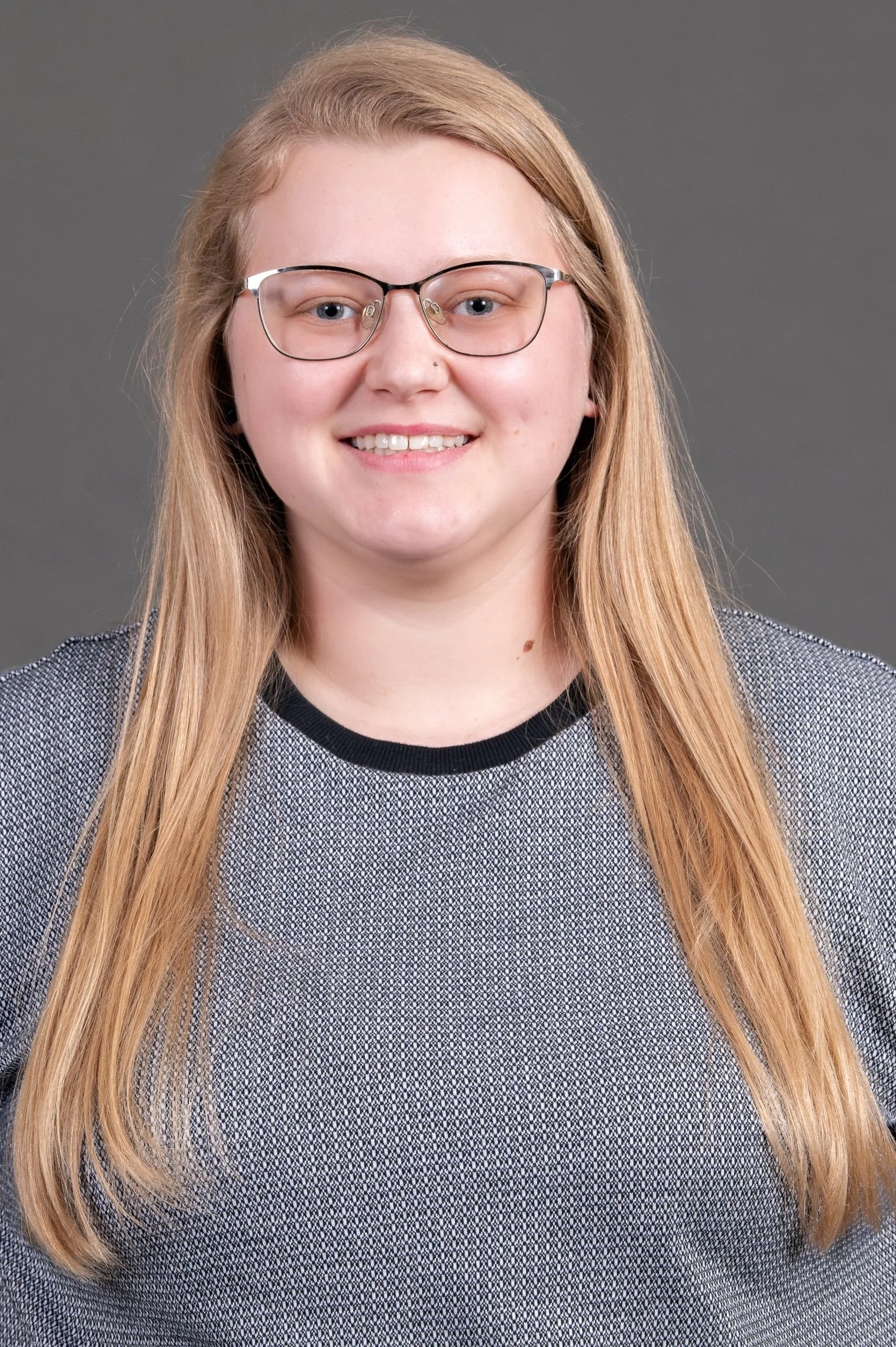A young woman with long blonde hair, glasses, and a nose piercing smiling at the camera against a gray background.