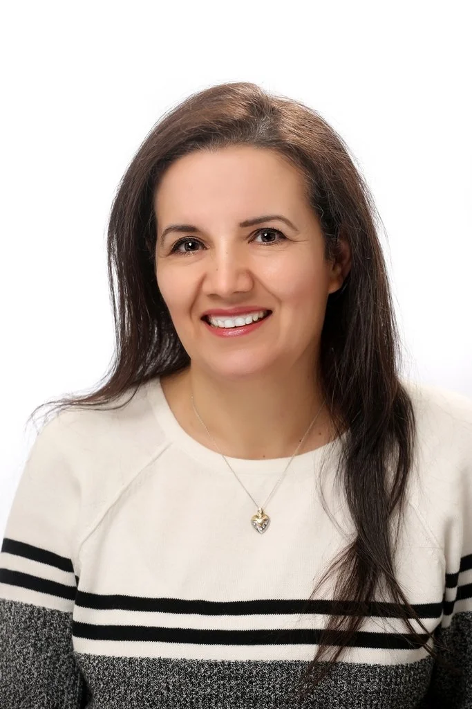 A woman with long brown hair, smiling, wearing a white sweater with black stripes and a heart-shaped pendant necklace, posing against a white background.
