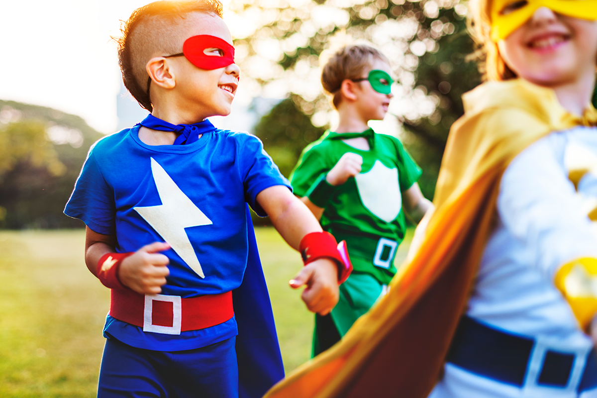 Three children dressed in superhero costumes running outdoors in a park.