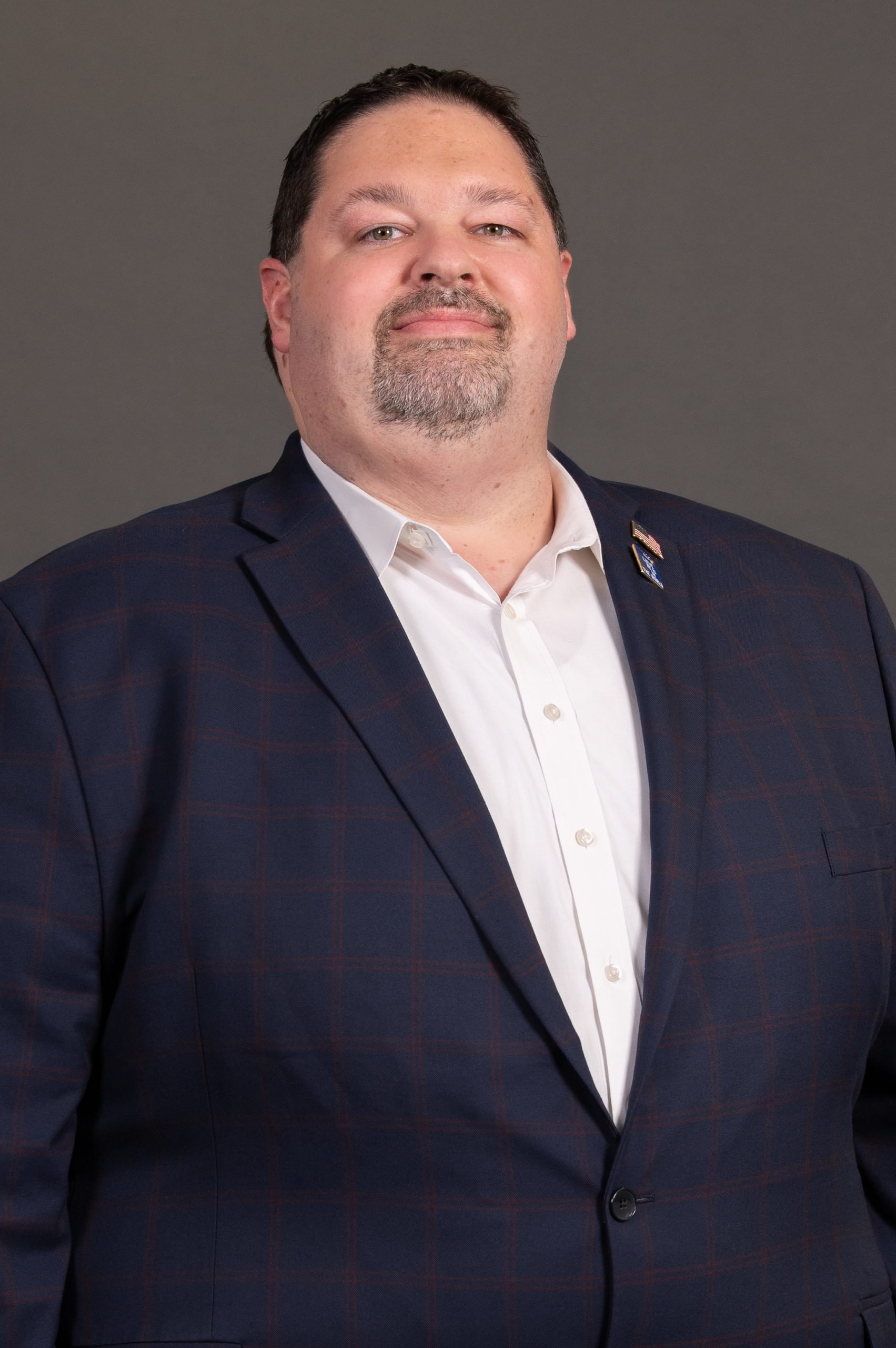 Portrait of a man in a suit with an American flag pin on his lapel, looking confidently at the camera against a gray background.