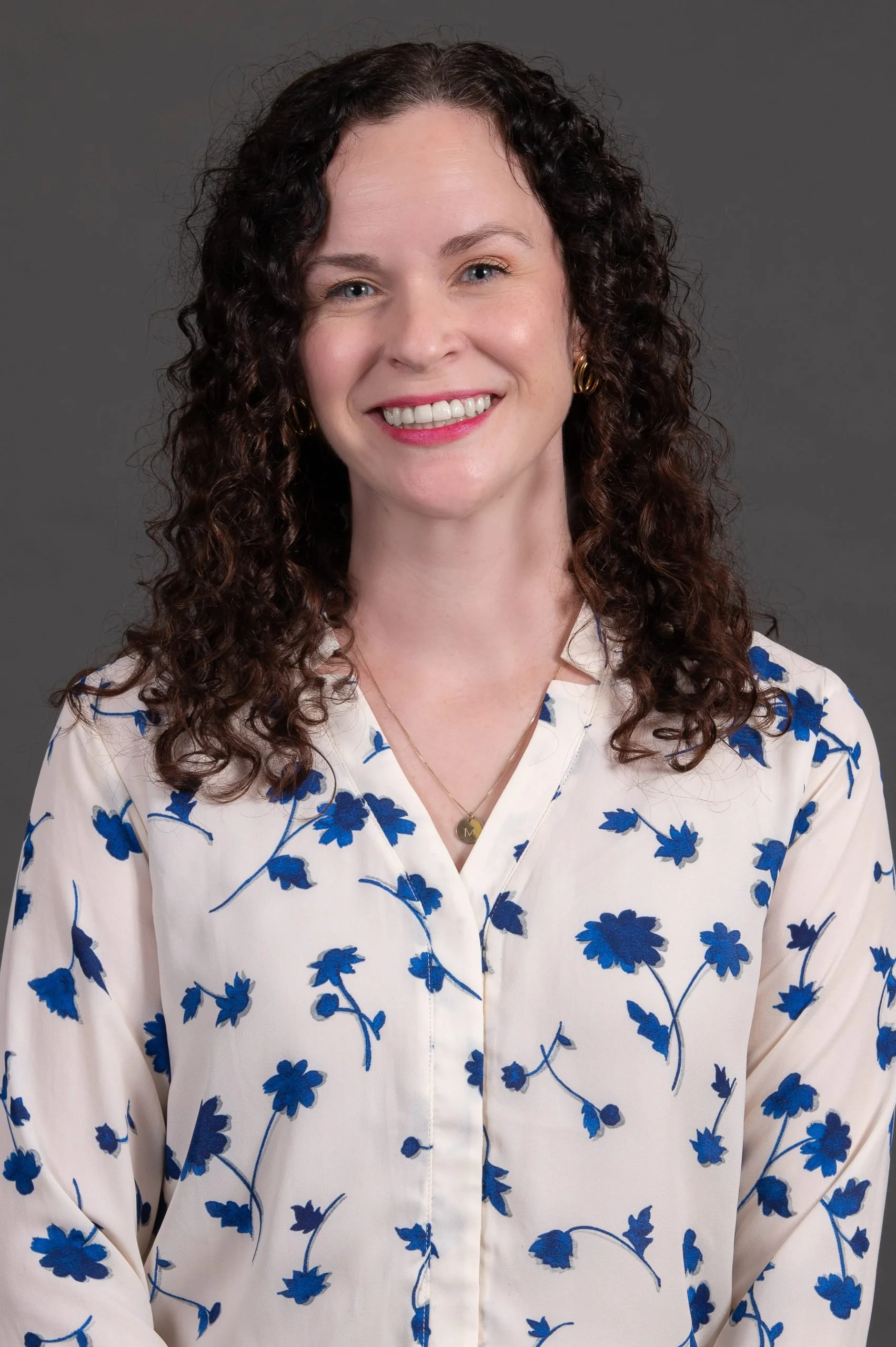 A woman with curly dark hair smiling, wearing a white blouse with blue floral prints, gold earrings, and a necklace with a round pendant, against a gray background.