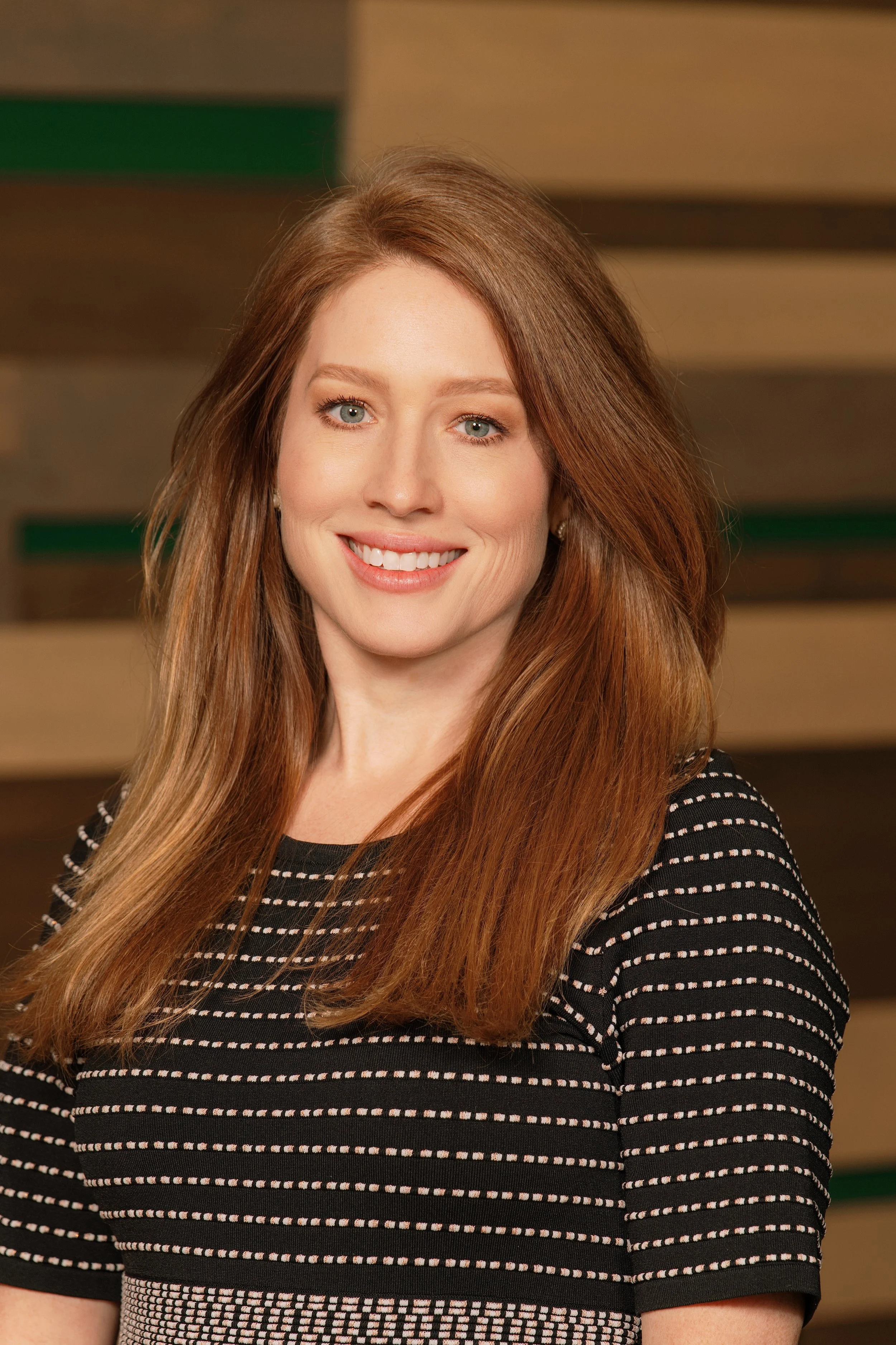 A woman with long red hair smiling, wearing a black and white striped top, standing in front of a background with wooden panels.
