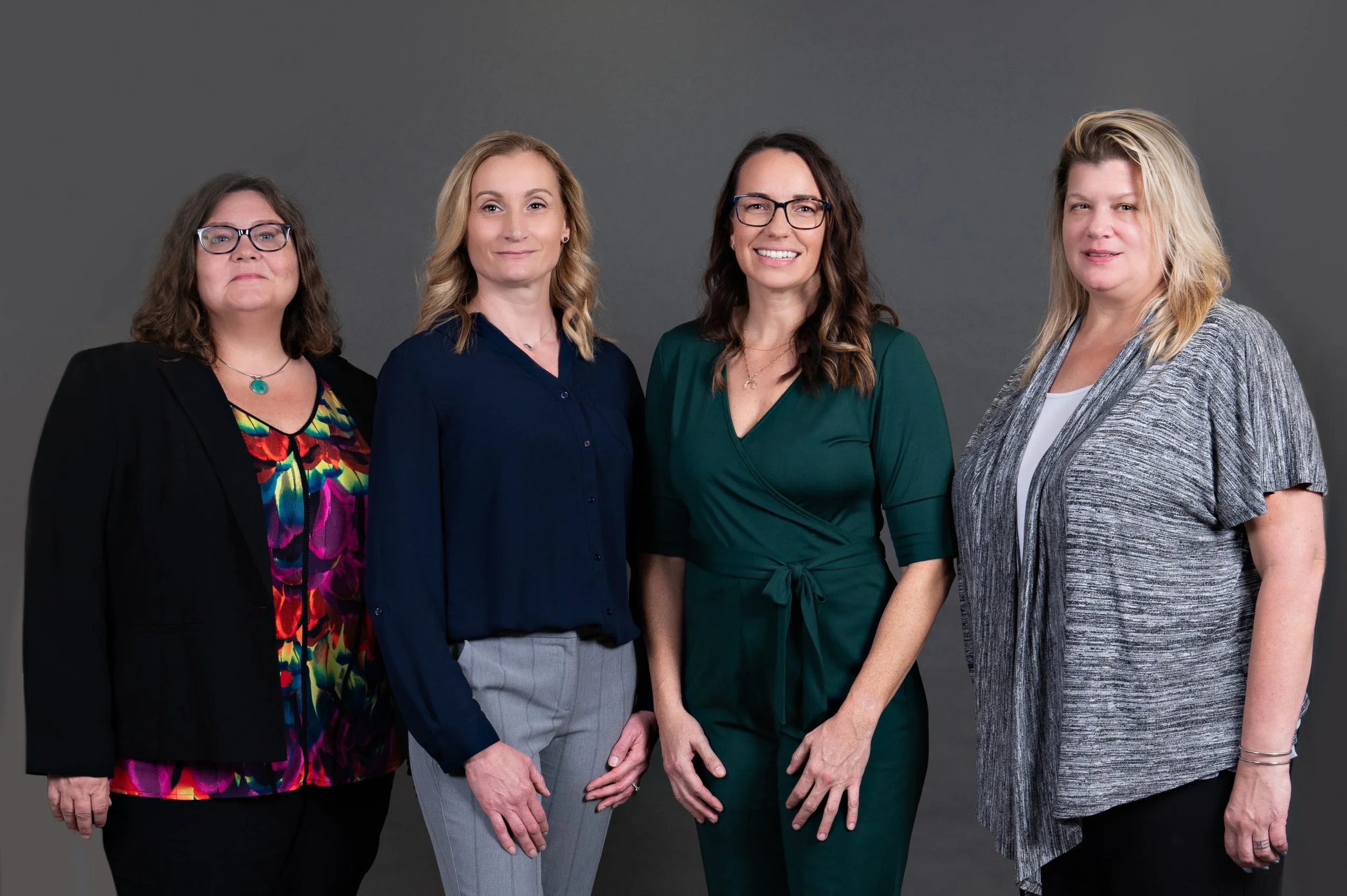 Four women standing side by side against a gray background, dressed in business casual attire.