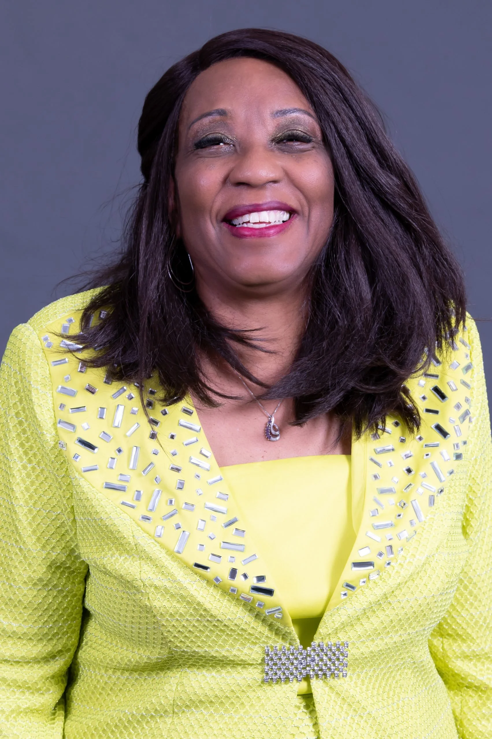 Smiling woman with dark hair wearing a yellow jacket with silver embellishments and a matching top, necklace, and earrings, against a gray background.