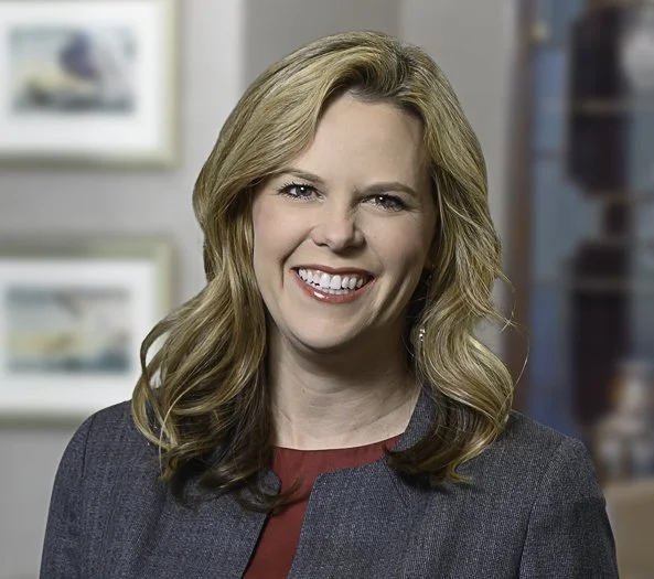 A woman with blond, wavy hair smiling in an indoor setting.
