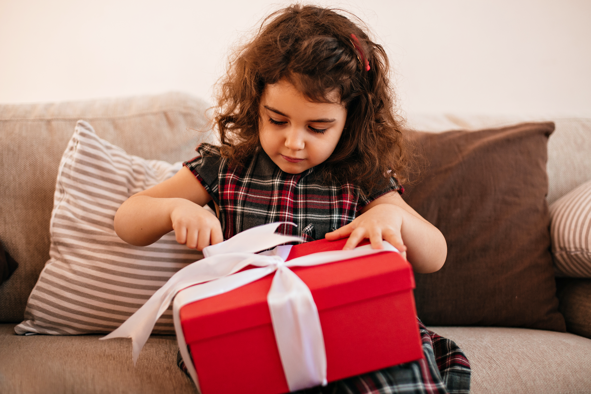 A young girl with curly hair and a red hair clip opening a red gift box with a white ribbon on a sofa with striped and solid pillows.