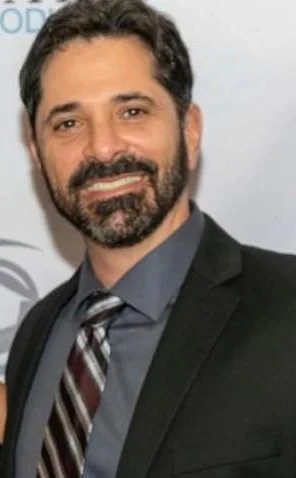 A man with dark hair and a beard wearing a dark suit, gray shirt, and striped tie, smiling at the camera.