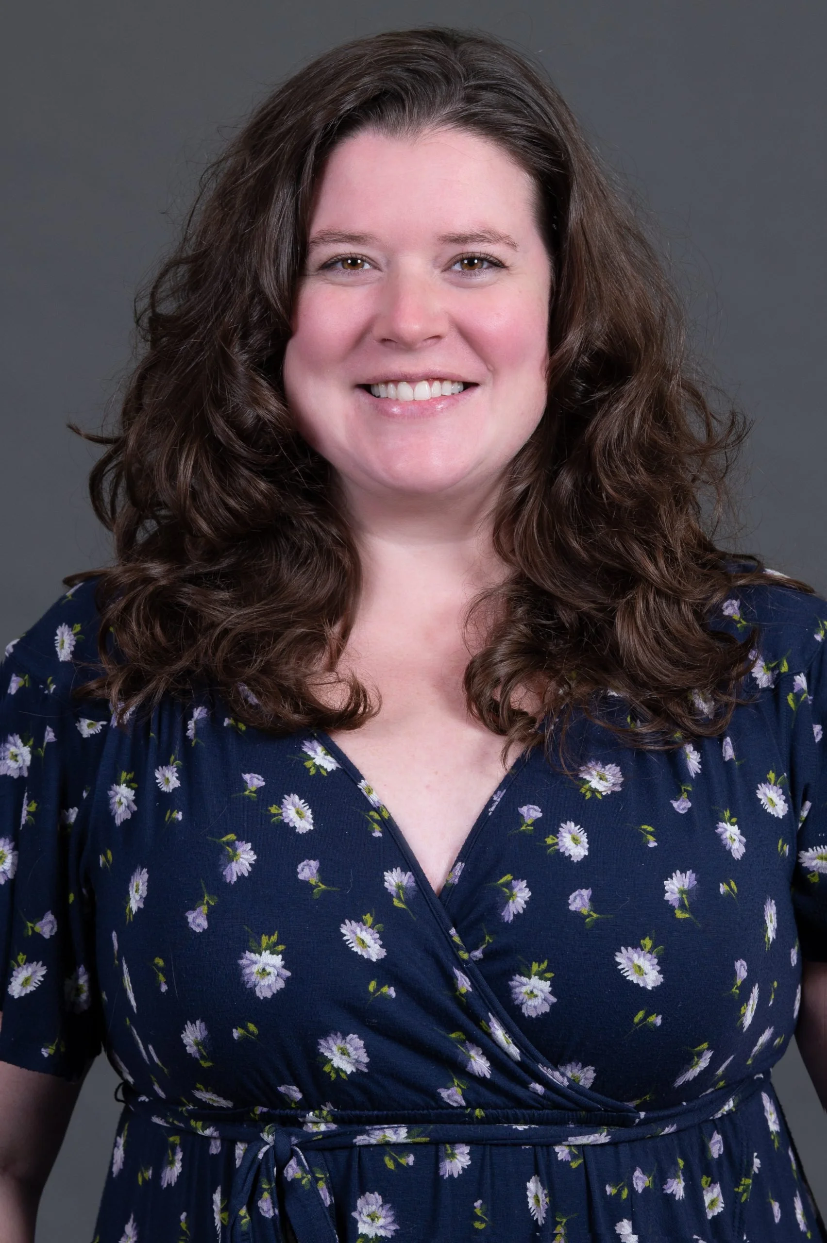 A woman with long, curly brown hair smiling at the camera, wearing a navy blue dress with a floral pattern, against a gray background.