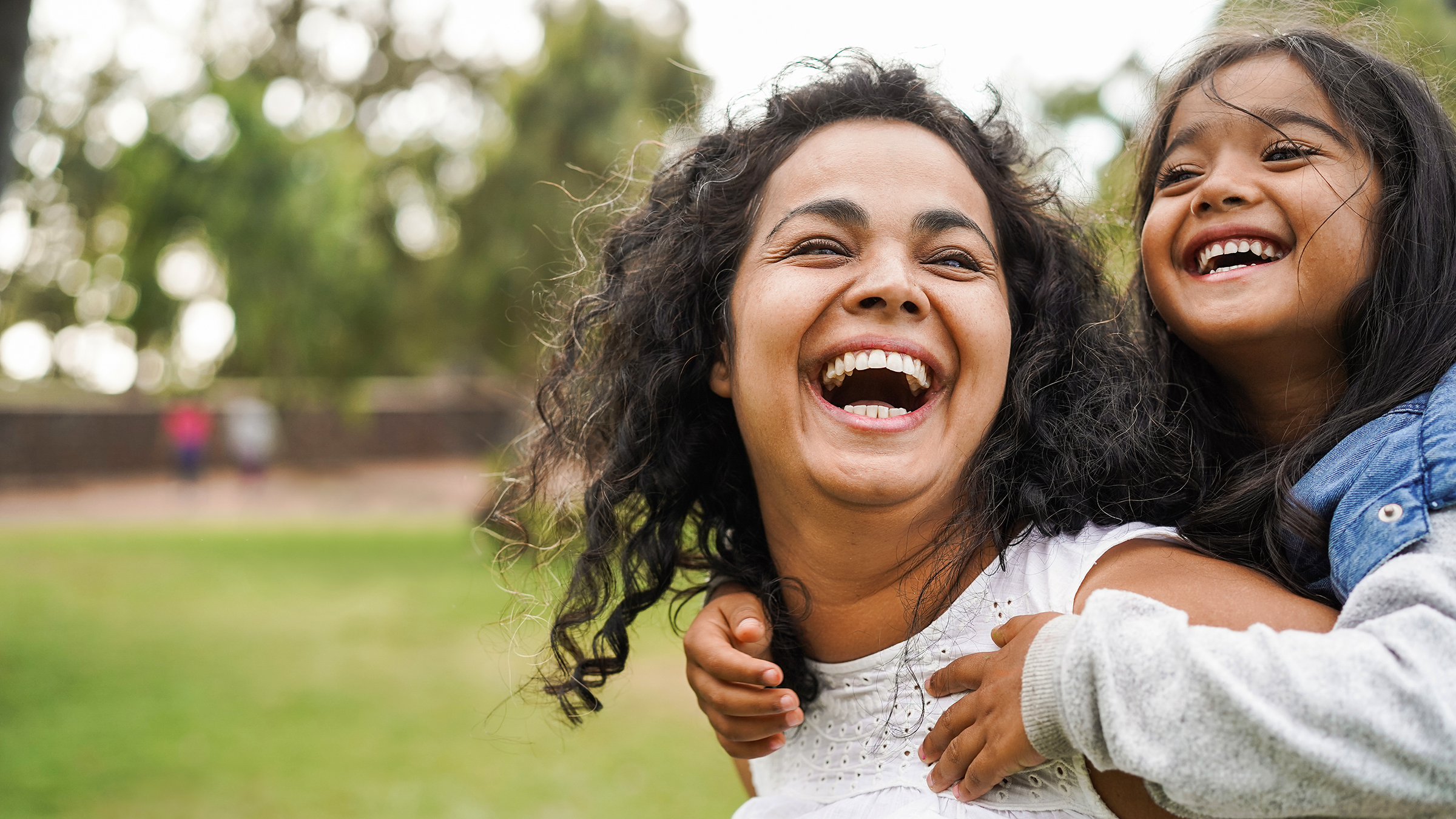 A woman giving a piggyback ride to a young girl outdoors in a park, both are laughing and smiling.