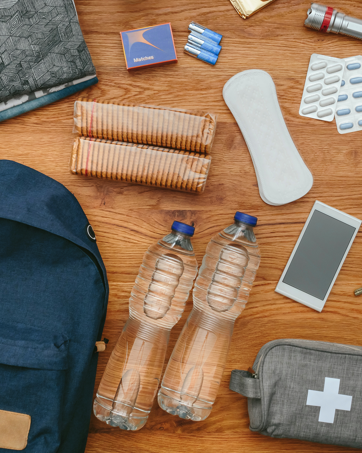 Items laid out on a wooden surface including bottles of water, a first-aid kit, a large menstrual pad, a flashlight, batteries, tissue packs, a smartphone, and a backpack.