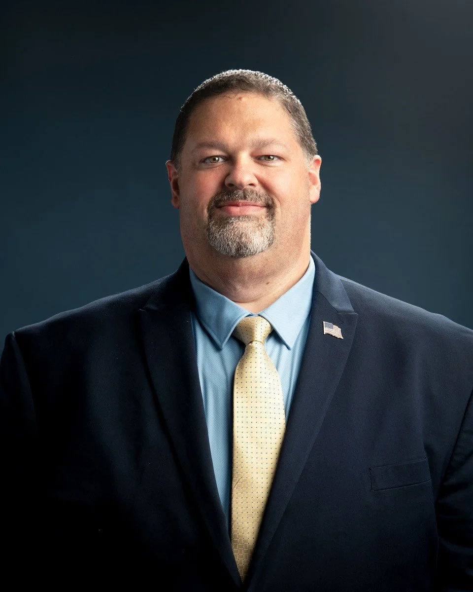 Portrait of a man in a suit with an American flag pin on his lapel, looking confidently at the camera against a gray background.