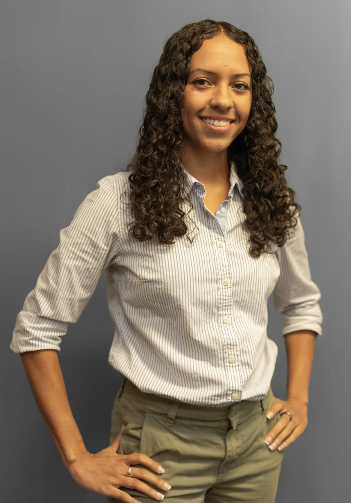 A woman with curly hair wearing a striped button-up shirt and khaki pants stands against a plain gray background smiling with hands on her hips.
