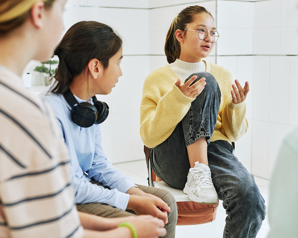 A young woman in glasses and a yellow sweater sits with one leg up on a chair and gestures as she talks to two other women during a conversation in a bright, white room.