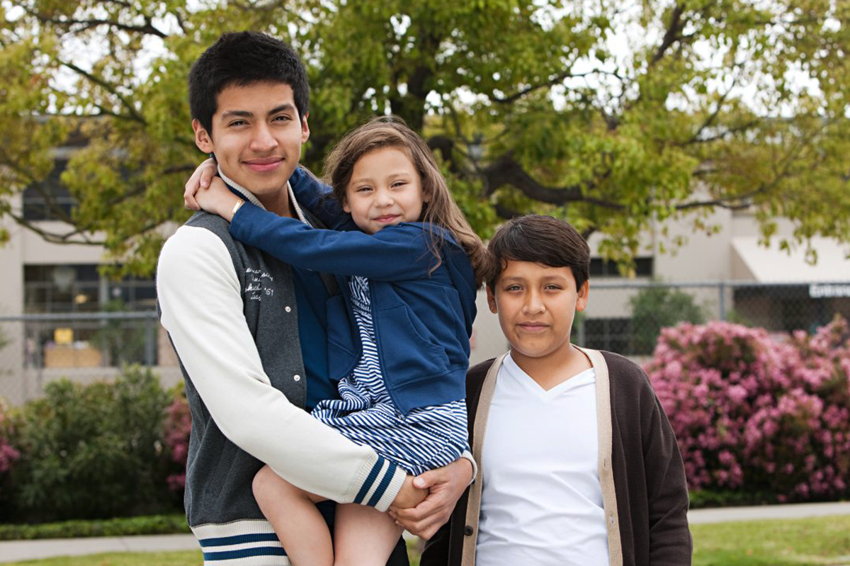 A young man holding a girl, with another boy standing next to them outdoors in front of a tree and some pink flowering bushes.