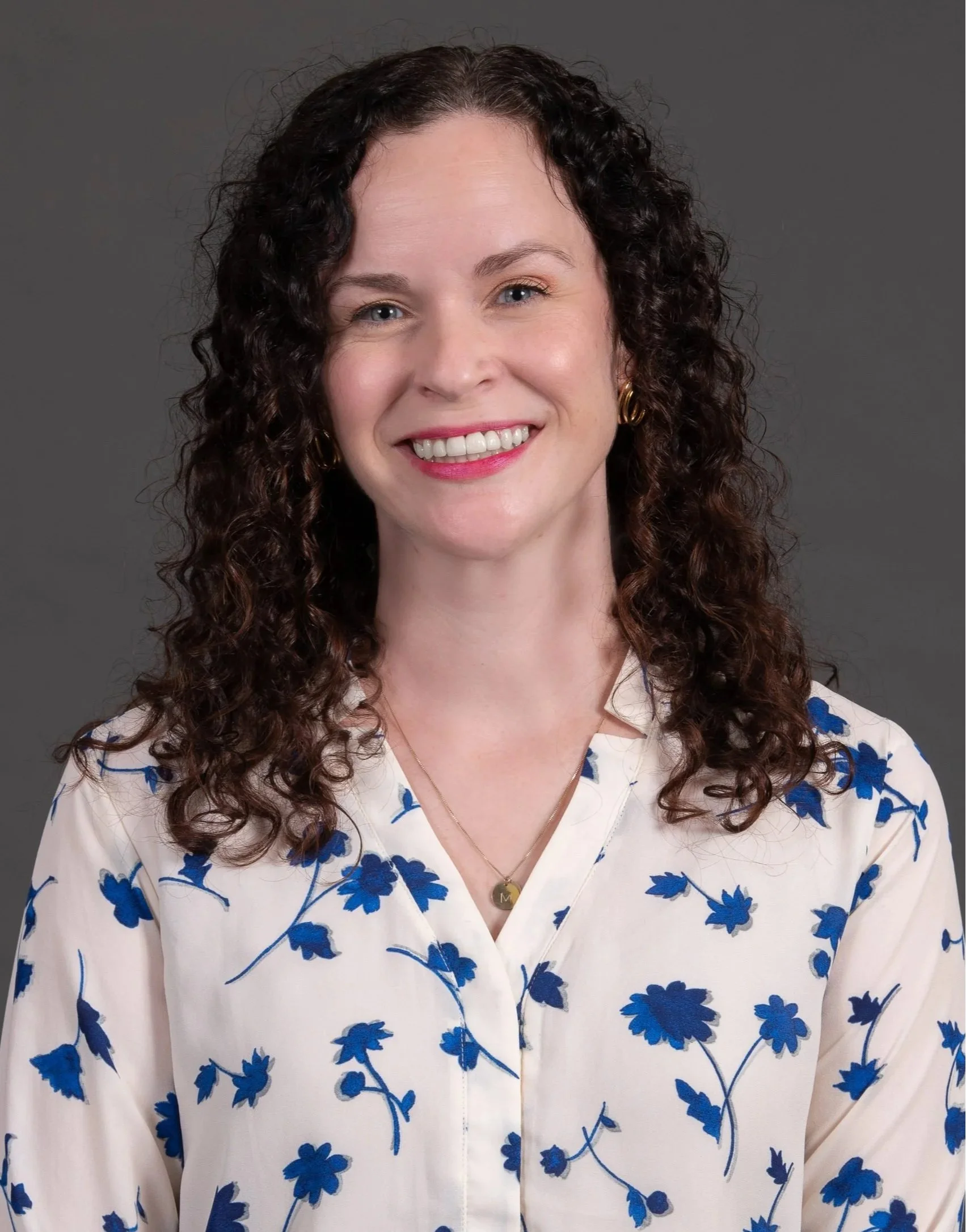 A woman with curly dark hair smiling, wearing a white blouse with blue floral prints, gold earrings, and a necklace with a round pendant, against a gray background.