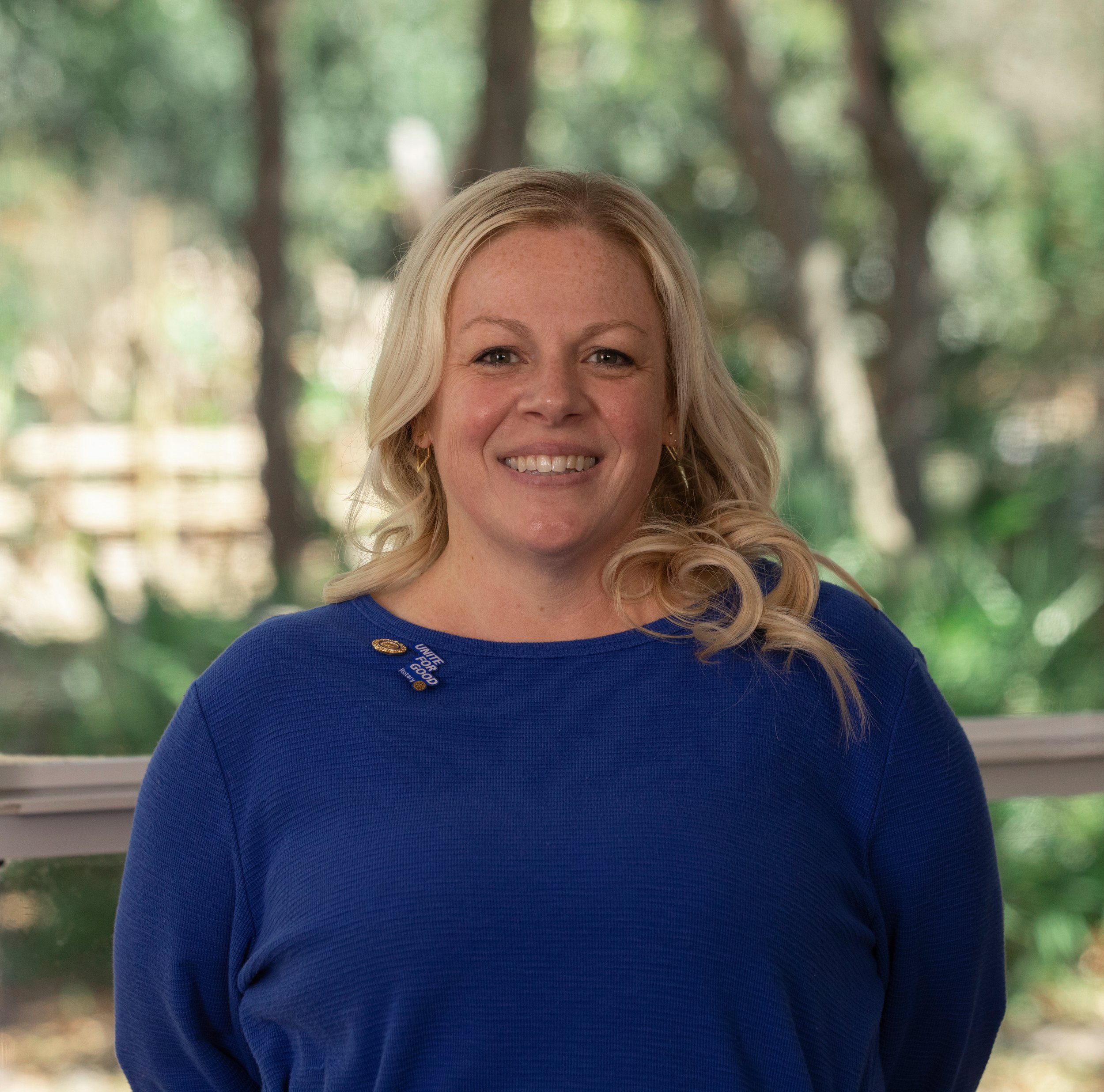 Close-up portrait of a smiling woman with blonde curly hair, wearing pearl earrings, outdoors with a bright background.