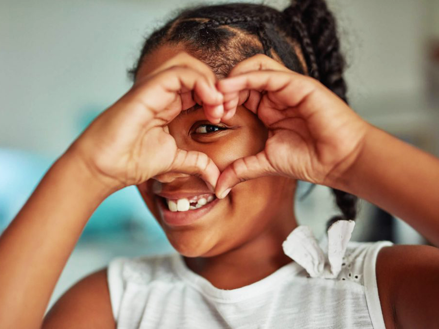 A smiling young girl with braids and a missing front tooth, making a heart shape with her hands over her face.